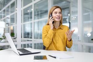 maduro exitoso rubia negocio mujer en amarillo camisa felizmente hablando en el teléfono con un sonrisa, hembra financiero trabajador a el lugar de trabajo en el medio de el oficina utilizando un ordenador portátil a trabajar. foto