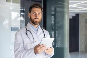 Portrait of a serious young male doctor standing in a white coat in the office of the clinic, holding a tablet in his hands and confidently looking at the camera. photo