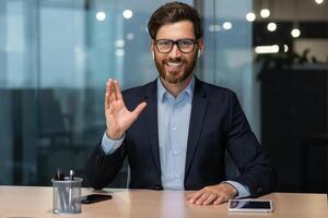 call on online meeting with colleagues, mature boss in business suit looking at web camera and smiling waving to employees, businessman in glasses working inside modern office building. photo