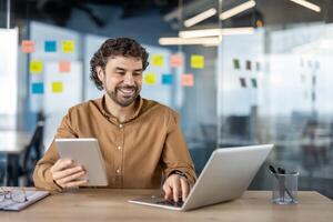 un alegre hombre en un casual camisa es multitarea con un tableta y ordenador portátil a un moderno lugar de trabajo con transparente pegajoso notas en el antecedentes. foto