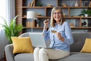 extático mujer a hogar haciendo un exitoso compra en línea con un crédito tarjeta y computadora portátil, expresando alegría y satisfacción. foto