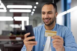 Confident young man holds a credit card and smartphone, shopping online with a smiling expression in a modern office setting. photo