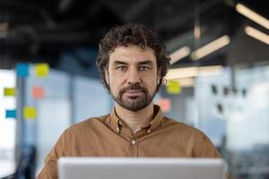 Portrait of a focused Hispanic businessman working on laptop computer and looking at camera in a modern office setting. photo