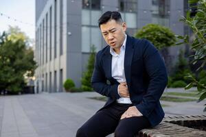 A man in a business suit appears distressed with chest pain, sitting outside on a bench in an urban setting. photo