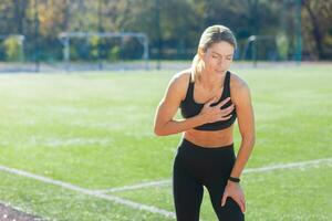 determinado mujer en ropa de deporte tomando un romper, atrapando su aliento post-entrenamiento en un soleado Deportes campo. foto