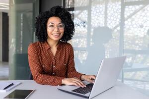 Portrait of mature adult successful businesswoman, african american woman at workplace smiling and looking at camera, confident boss working at workstation inside office, using laptop. photo