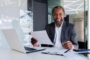 Portrait of successful mature experienced financier, business man behind paper work smiling and looking at camera, african american man at workplace reviewing contracts, accounts and reports. photo