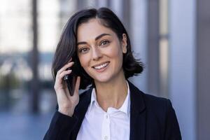 de cerca retrato de un joven hermosa mujer de negocios en pie en un traje en el calle, hablando en el teléfono y mirando a el cámara. foto