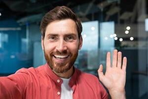 Young happy businessman looking into smartphone camera and talking on call, man working inside modern office building, smiling and talking remotely with friends, using communication app. photo