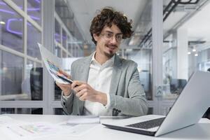 un joven, experimentado empresario con Rizado pelo atentamente examina gráficos mientras trabajando en un brillante oficina ambiente. foto