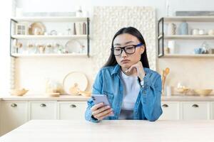 triste mujer a hogar sentado en cocina leyendo Noticias en teléfono, Deprimido asiático mujer participación teléfono inteligente en lentes y camisa. foto