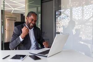 Mature experienced businessman celebrating victory success at workplace, boss inside office happy with financial results achievement received online notification win message, holding hands up. photo