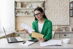 un mujer trabajando a hogar con un ordenador portátil. ella es participación un sobre con un letra en su manos foto