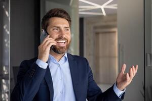 Close-up photo of a smiling young businessman standing inside an office and talking on the phone with clients and partners, gesturing with his hands.