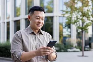 Close-up photo of a smiling young Asian man, an office worker standing outside and using the phone.