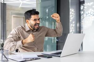 exitoso empresario dentro oficina celebrando victoria y triunfo, hombre leyendo contento Noticias desde computadora portátil, empresario trabajando a lugar de trabajo, satisfecho con logro resultados. foto