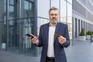 Angry businessman holding a card and a smartphone, standing near an office building, unsuccessful online shopping, fraud on the Internet. photo