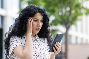 Sad disappointed woman received online notification with bad news on her phone, businesswoman walking outside office building, using application on smartphone, reading social media unsatisfied. photo