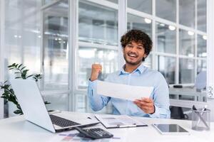 Portrait of successful hispanic businessman, man in shirt reading report and happy celebrating success satisfied with result holding hand up, worker inside office with laptop working on paper work. photo