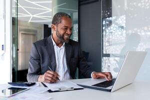 exitoso satisfecho financiero detrás papel trabajar, afro americano mayor experimentado relleno fuera documentos, utilizando calculadora para cálculo, empresario detrás papel trabajo dentro oficina sonriente. foto