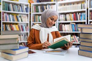 joven hermosa hembra estudiante estudiando en el biblioteca, hembra estudiante en hijab sonriente entre libros estudiando material independientemente leyendo literatura. foto