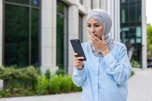 joven negocio mujer en hijab caminando en el ciudad, árabe mujer recibido en línea notificación con malo noticias, participación teléfono en manos leyendo trastornado y Deprimido triste. foto