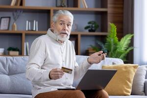 A senior Caucasian man sits on a couch, holding a credit card and looking confused while using a laptop. The setting is a cozy, modern living room. photo