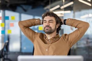 Calm caucasian guy sitting on chair with hands behind head and closed eyes in background of stylish office. Digital designer daydreaming and setting mind up for creative mood at beginning of workday. photo