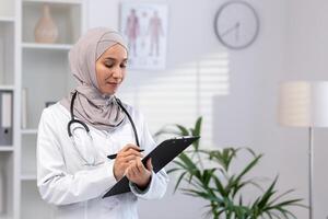 un dedicado hembra musulmán médico en un hijab y blanco Saco es enfocado en pacientes informes mientras trabajando en un clínica. foto