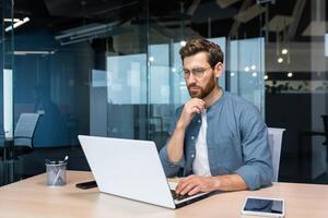 .grave pensativo empresario en camisa pensando acerca de decisión sentado a mesa en moderno oficina, hombre con barba es utilizando ordenador portátil a trabajar. foto