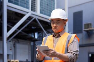 Close-up photo of a young Asian man in a hard hat and vest standing outside a building and factory and using a tablet, in concentration.