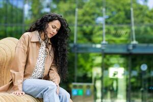 Tired caucasian woman with long black curly hair sitting on bench outdoors touching and experiencing discomfort in knee. Background of part of glass structure or building. photo
