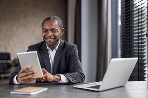 A cheerful professional black businessman in a suit sits at a modern office desk, happily using his tablet with a laptop nearby. photo