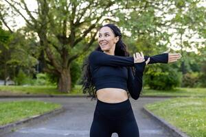 enfocado mujer en ropa de deporte ejecutando un brazo estirar, preparando para un rutina de ejercicio en un verde parque ajuste. foto