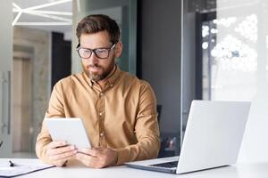 grave concentrado hombre trabajando dentro oficina con tableta computadora, pensando maduro empresario utilizando computadora portátil, leyendo datos, y comprobación en línea reporte financiero datos. foto