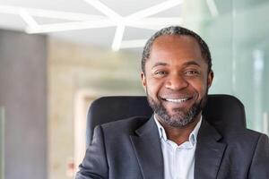 Close up portrait of successful successful businessman, man looking joyfully at camera sitting at desk inside office, satisfied african american boss. photo