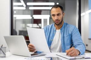 enfocado indio empresario revisando documentos mientras trabajando remotamente desde un contemporáneo vivo habitación, exudando profesionalismo y dedicación. foto