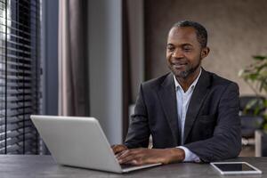 Grown up stylish man using portable gadgets for work in spacious apartment with window and plants. Progressive manager setting workspace at home and getting along with modern technology. photo