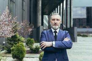 retrato de un mayor canoso hombre director, CEO en el antecedentes de el oficina centrar y arboles él es en pie grave en un traje con atar, brazos cruzado en frente, mirando a el lado. foto