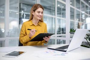 Successful mature female financial accountant at workplace inside office, fills data in table, works with documents and papers, uses laptop and calculator for paperwork. photo