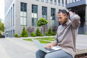Serious and pensive man sitting on bench unhappy with work result, using laptop for remote work, african american businessman outside office building sitting on bench photo