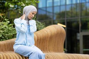 Middle-aged woman in a blue hijab experiences neck pain. She sits on a bench outdoors, holding her neck in discomfort near a modern building. photo