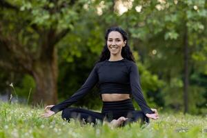 A serene young woman in black sportswear meditating in a lotus pose amidst vibrant green foliage and a calm, natural setting. photo