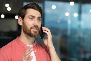 exitoso empresario en un rojo camisa hablando en el teléfono dentro el oficina, un hombre con un barba a el lugar de trabajo es hablando a un cliente, un satisfecho jefe en casual ropa. foto