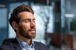 Mature businessman boss with eyes closed meditating and breathing in office, man with beard and business suit dreaming and visualizing inspiration. photo