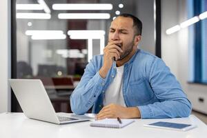 A tired young Muslim man in a blue shirt sits at a desk in a modern office and yawns while covering his mouth with his hand. photo