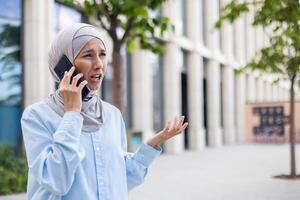 trastornado y enojado negocio mujer hablando en el teléfono, musulmán mujer en hijab infeliz con logro resultados caminando en el ciudad fuera de oficina edificio. foto