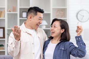 un joven asiático pareja, un hombre y un mujer, estar abrazando en un casa, departamento, y sostener llaves en su manos. sonriente personas Mira a cada otro, contento compradores, alquilar. foto