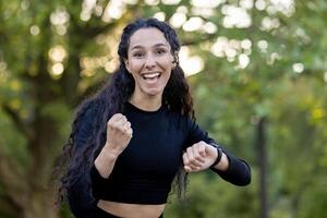 Joyful hispanic woman with fists pumped in victory pose outdoors, expressing excitement and happiness in a natural park setting. photo