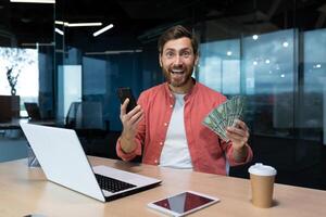 retrato de ganador hombre a lugar de trabajo dentro oficina, empresario mirando contentamente a cámara, participación teléfono y efectivo dólares, utilizando en línea aplicación en teléfono inteligente foto